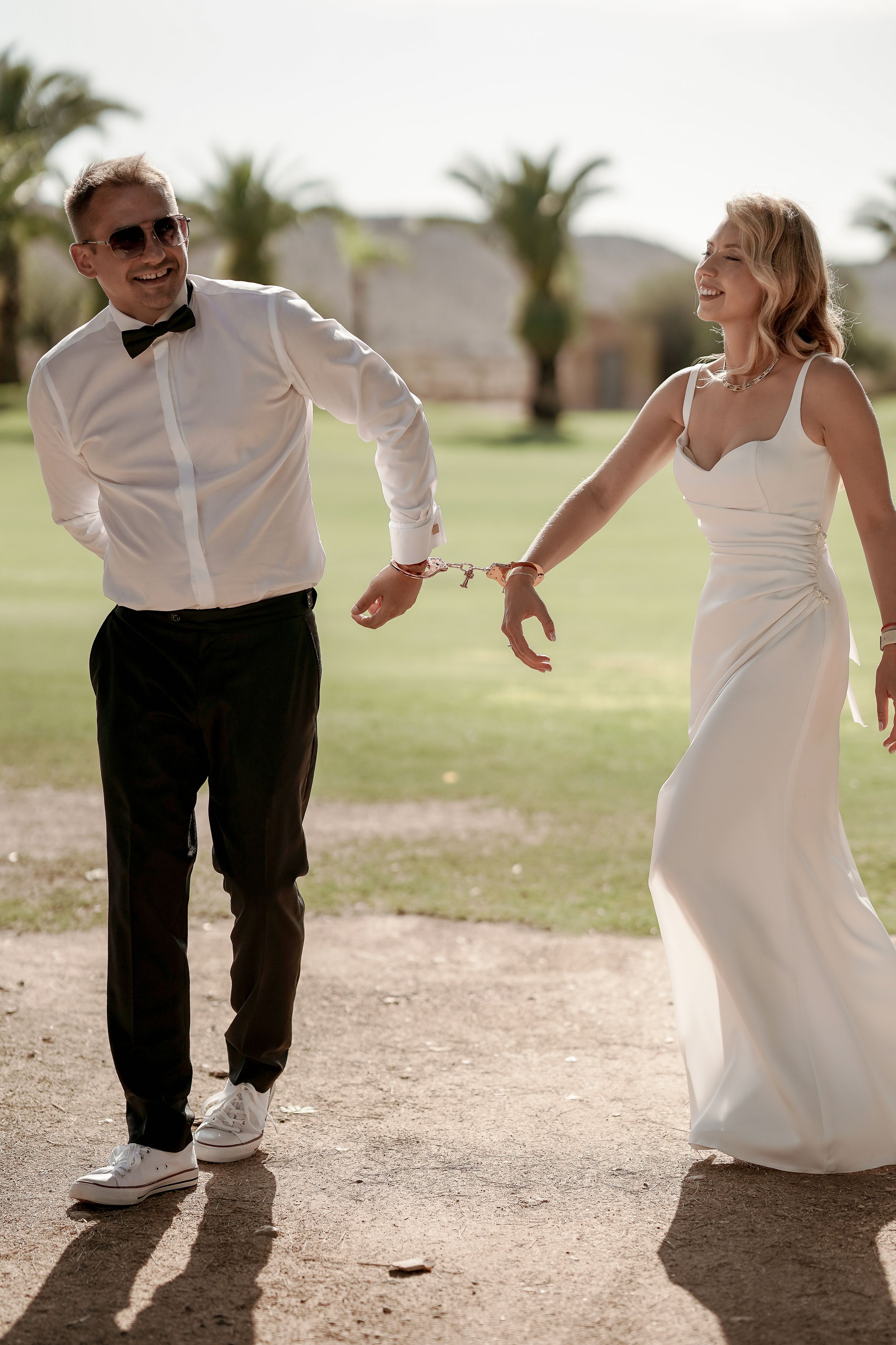 A couple in wedding attire playfully runs and dances together on a dirt path outdoors.