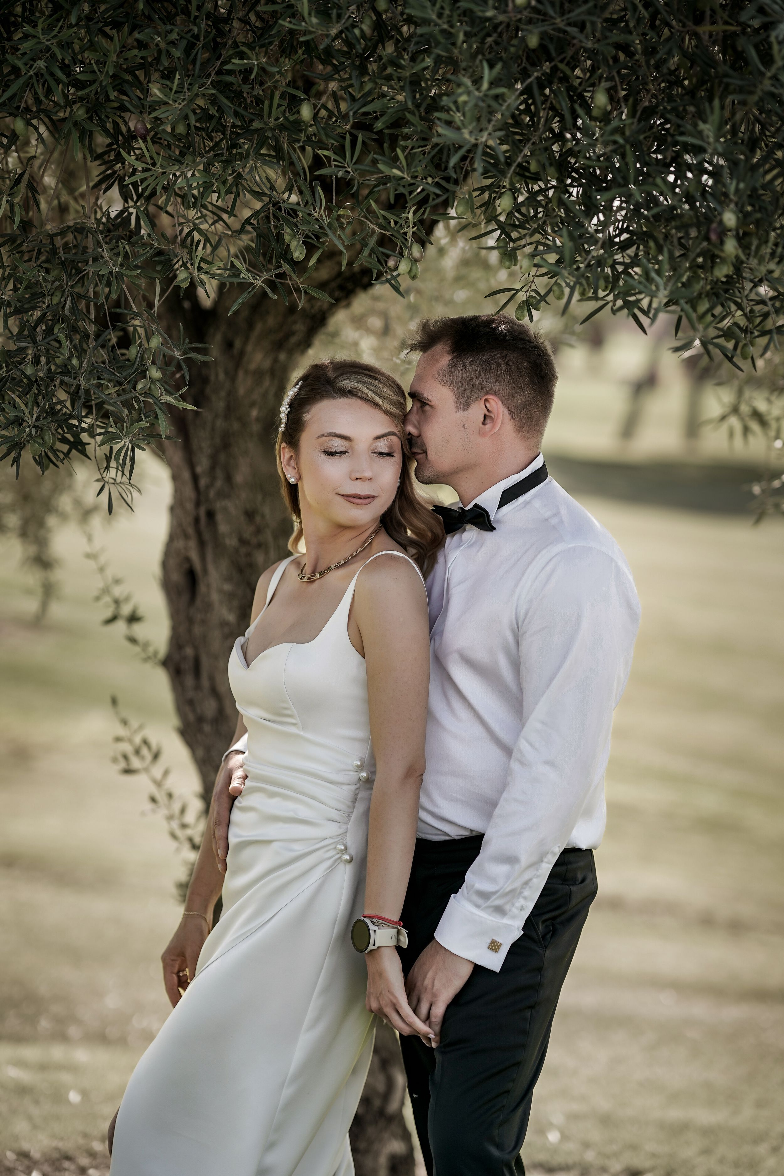 A romantic wedding couple embraces under olive trees in a sun-dappled garden setting.