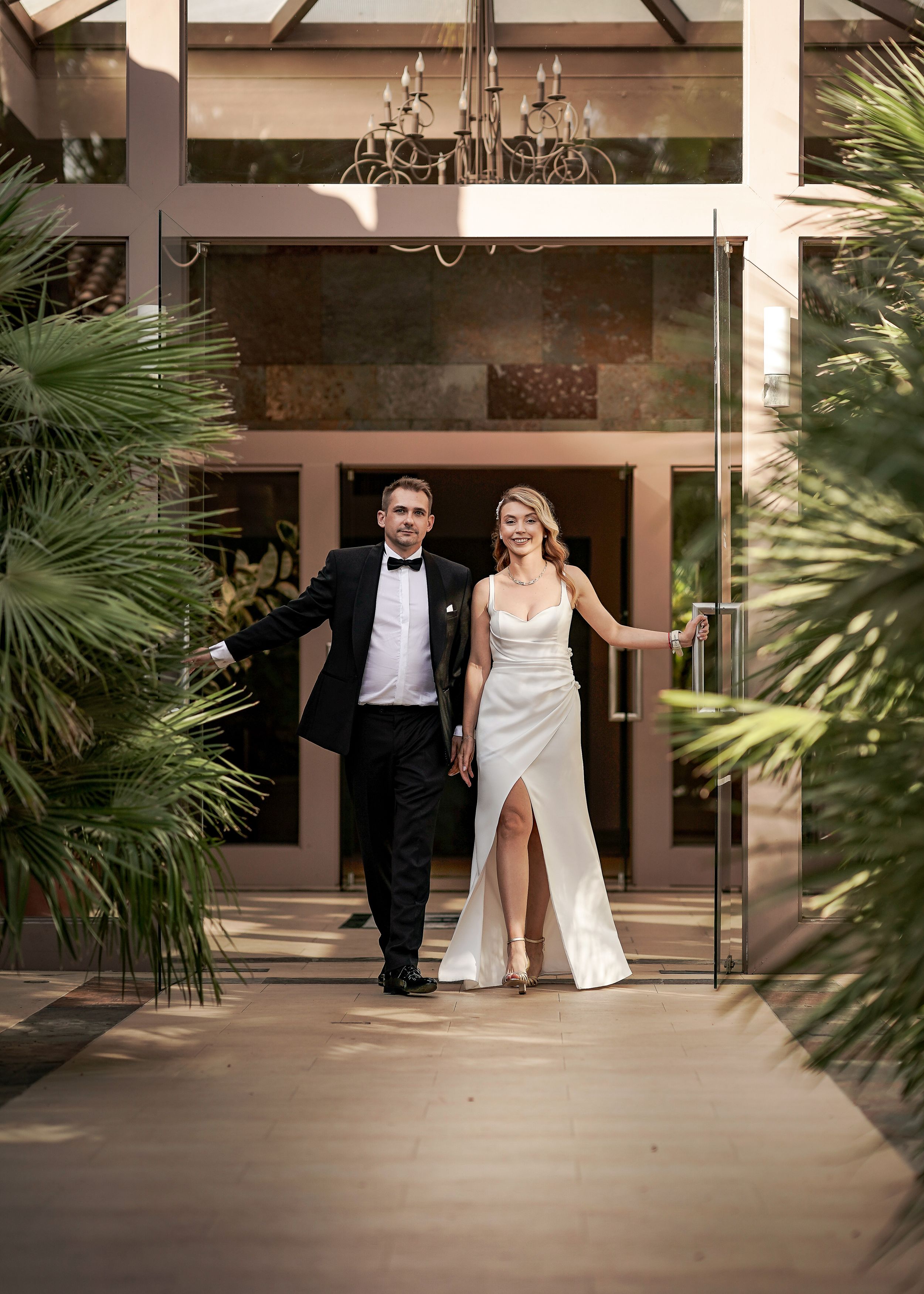 A newlywed couple exits through a tropical palm-lined walkway with an elegant chandelier overhead.
