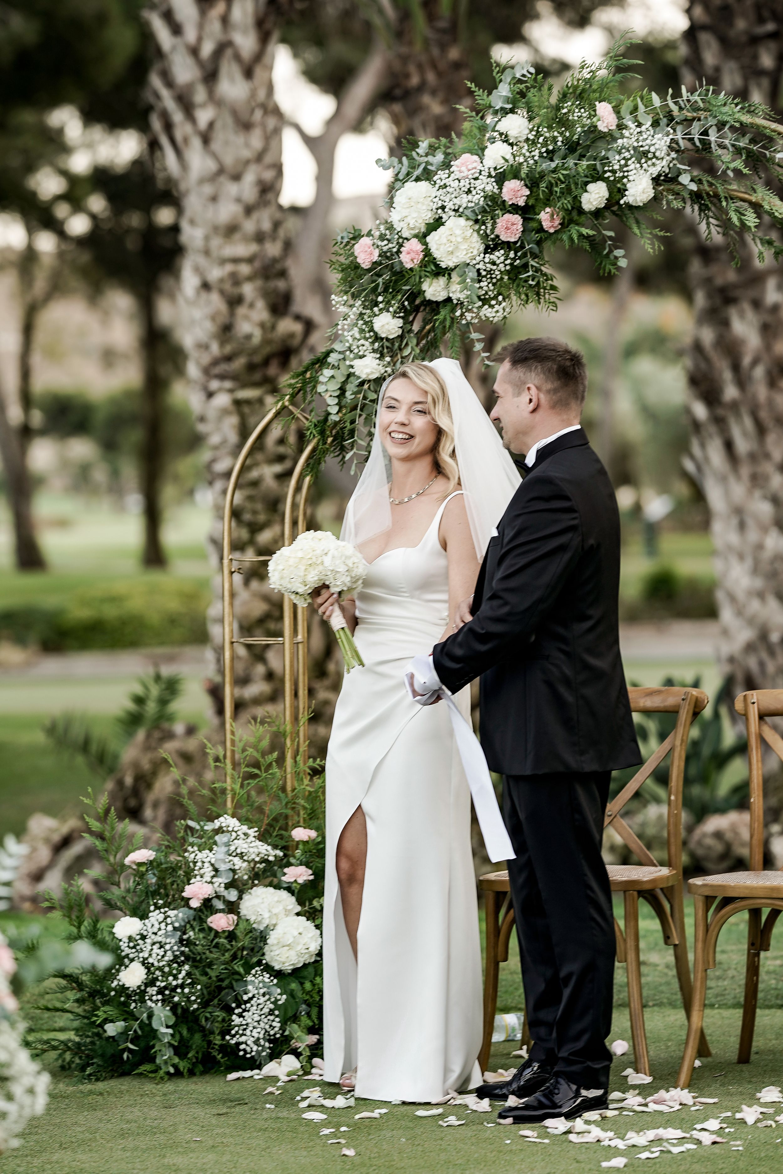Elegant outdoor wedding ceremony with floral decorations and wooden chairs arranged on a grassy lawn beneath birch trees.