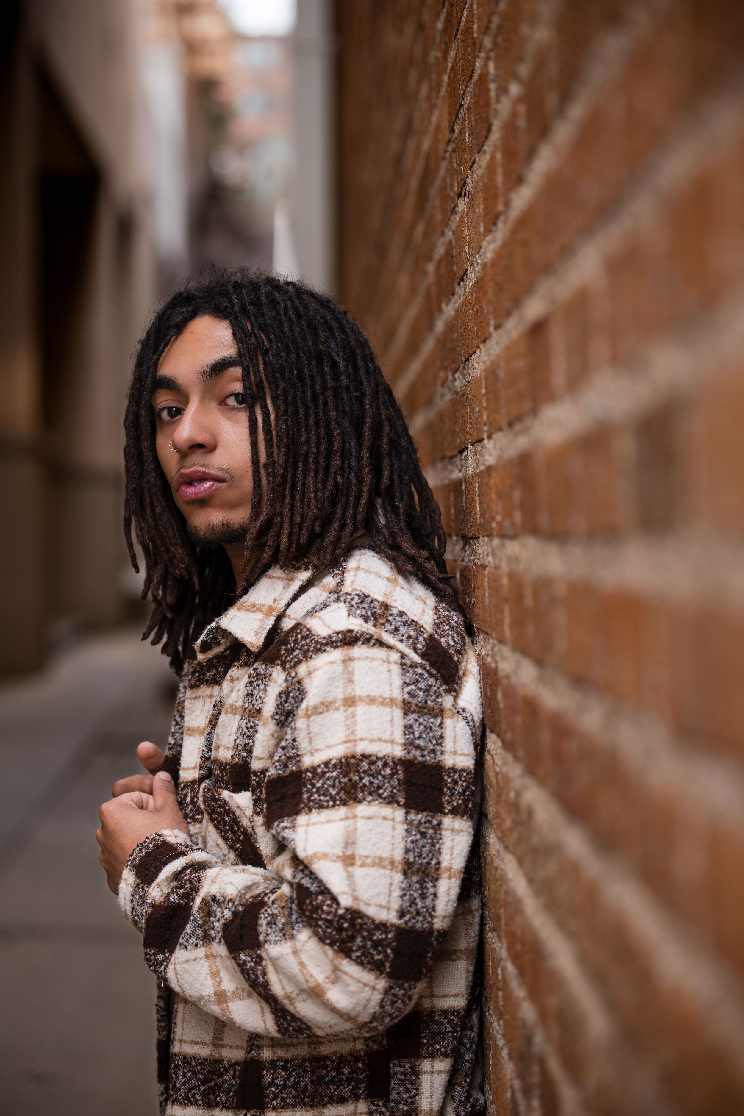 Artistic portrait against textured brick wall featuring plaid jacket and dreadlocks.