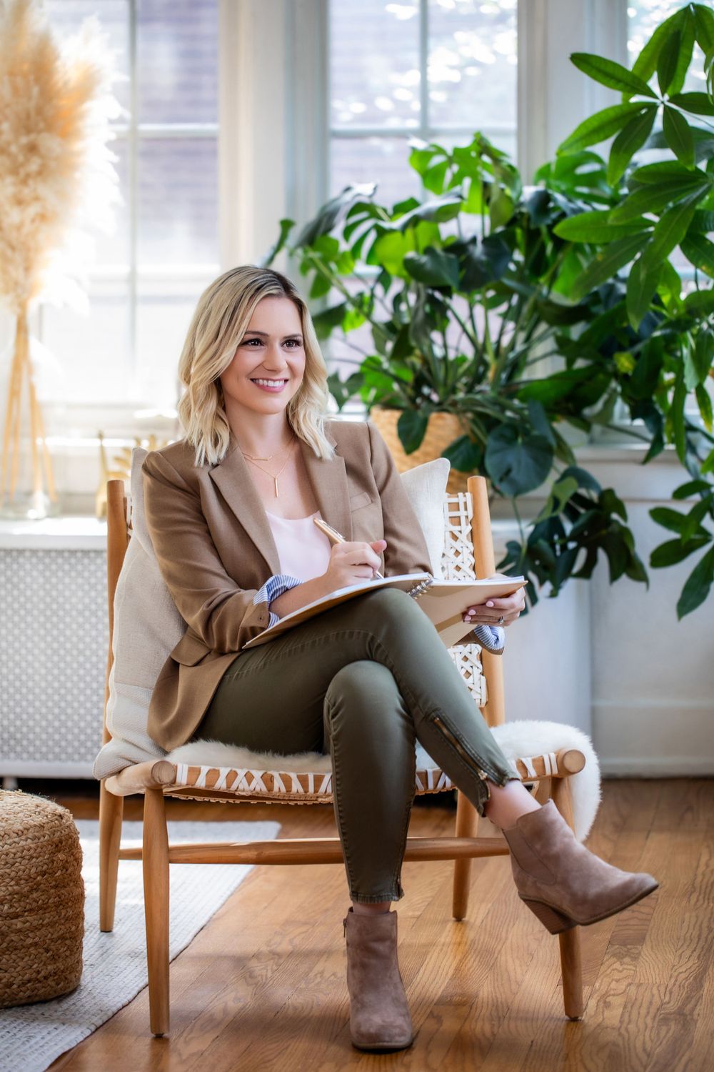 Person in beige blazer taking notes while sitting in woven chair near houseplants.