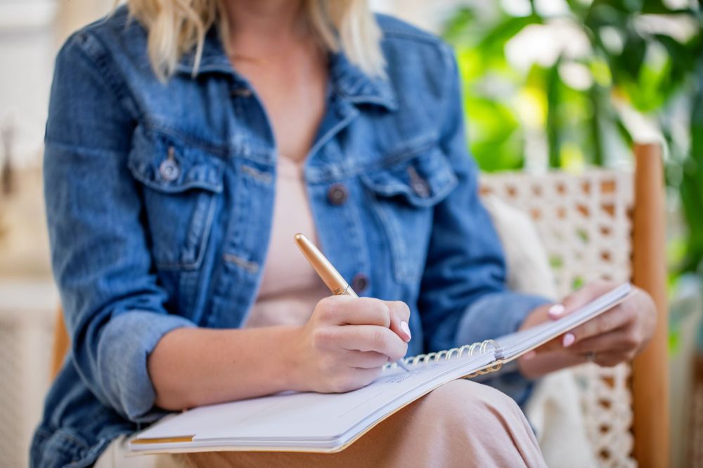 Person in denim jacket writing in spiral notebook while sitting outdoors.