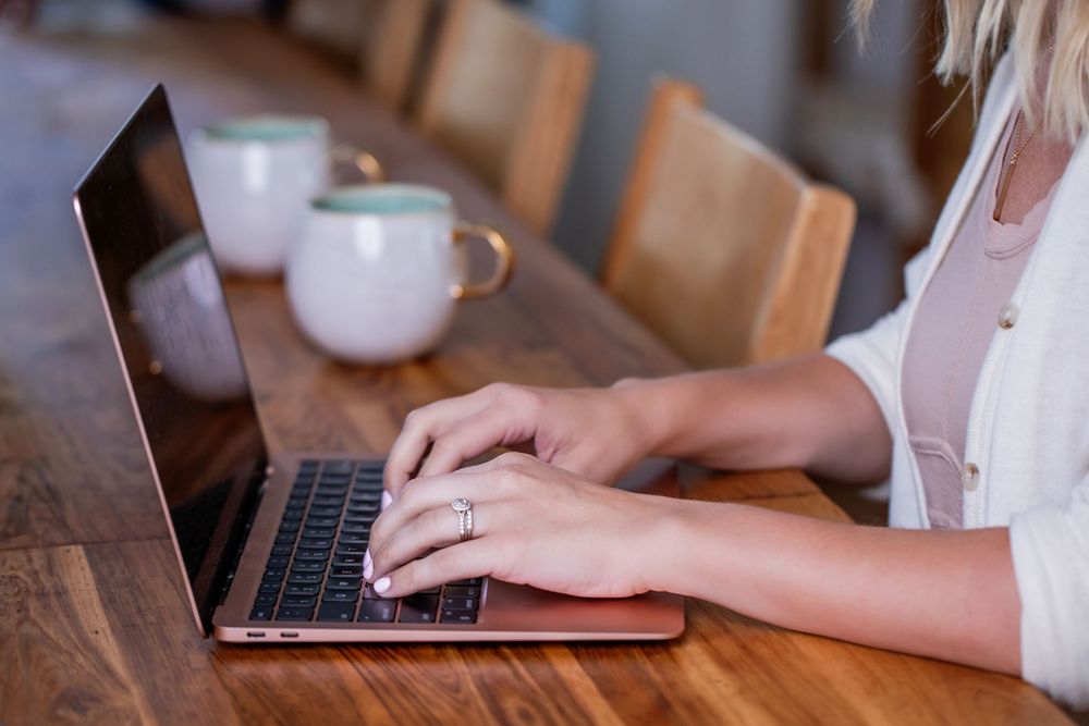 Hands typing on laptop keyboard with white coffee mugs on wooden table.
