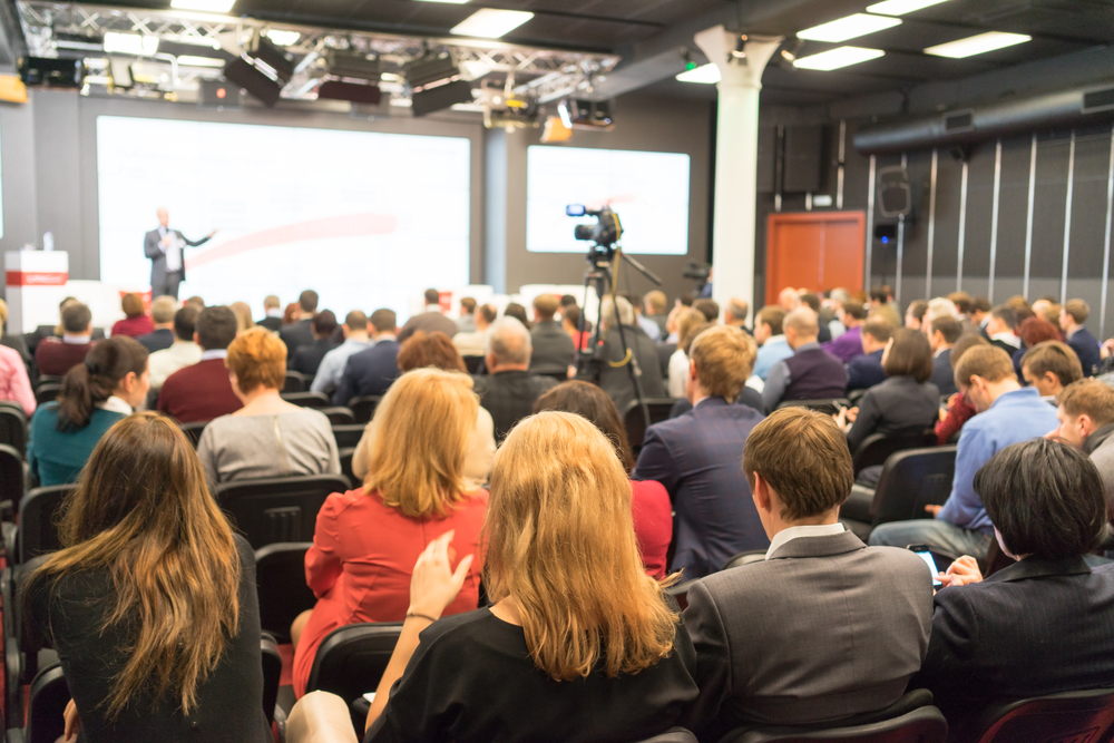 Large audience seated in rows at a business conference or seminar with video cameras and stage visible at front.