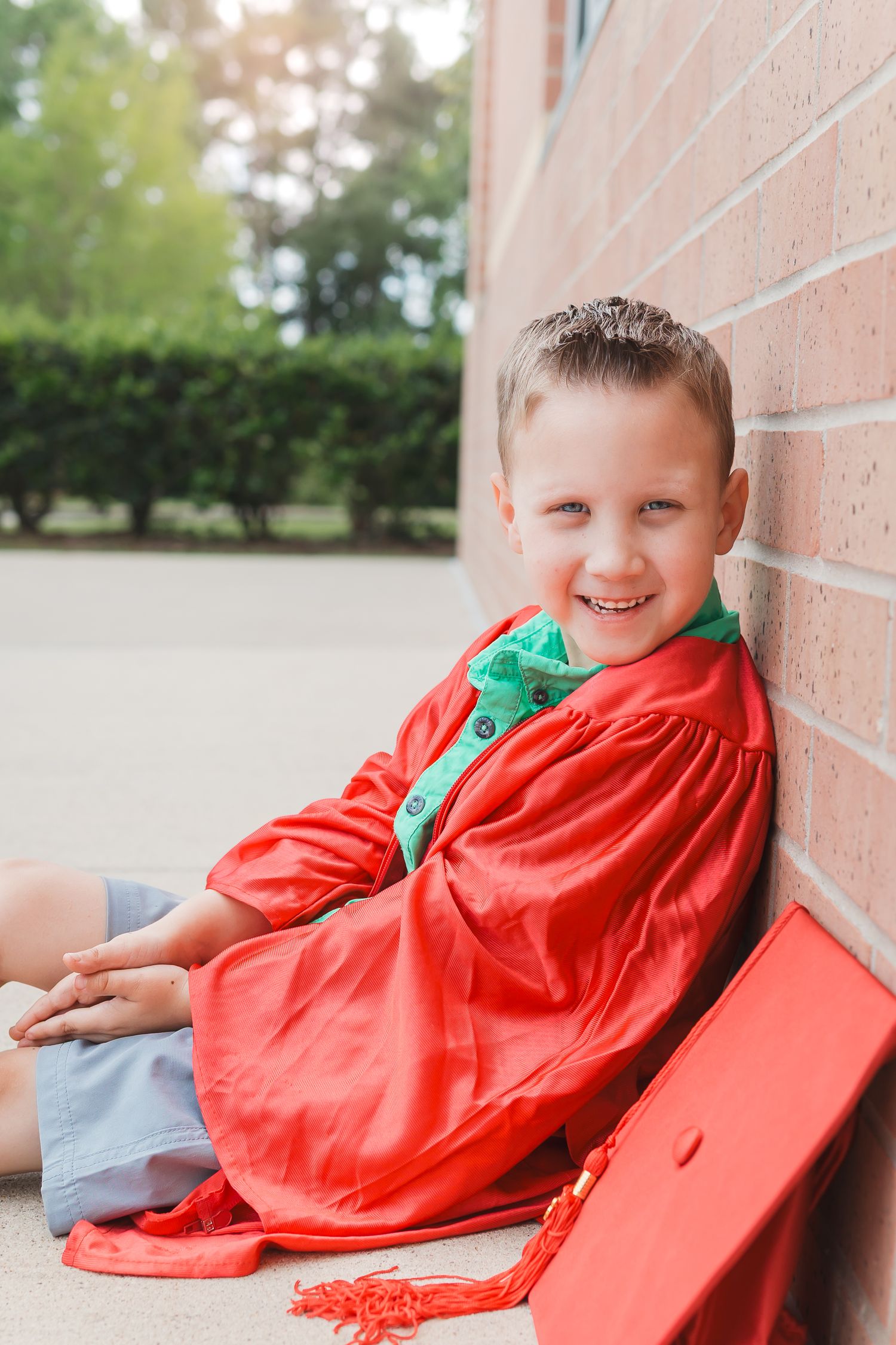 Young boy in red graduation cap and gown standing in green field.