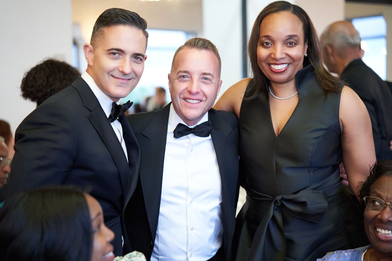 Three elegantly dressed guests smile together at a formal event, wearing black tie attire.