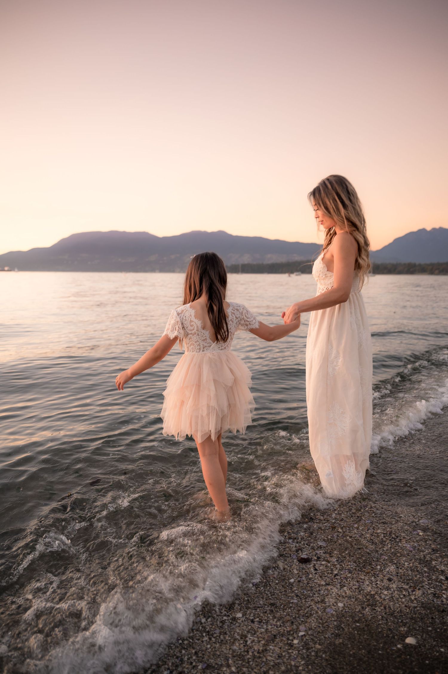 A romantic couple embraces on a sandy beach at sunset with calm ocean waters and mountains in the background.