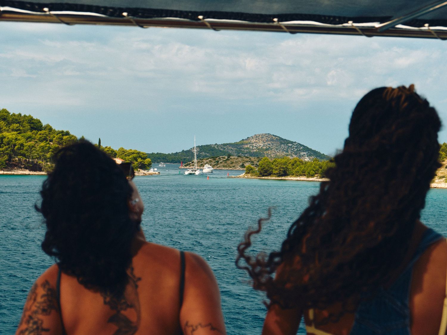 Two people enjoying a coastal view with sailboats and a mountain in the background on a sunny day.