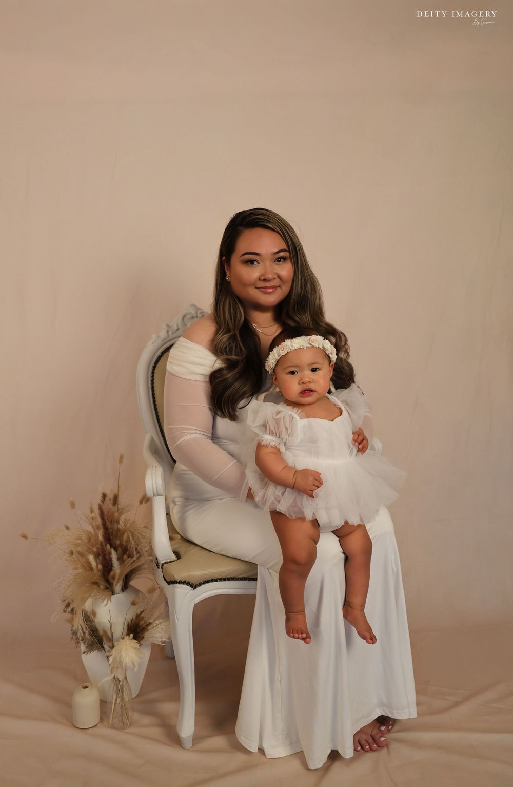 Mother and baby in matching white dresses pose for elegant studio portraits with neutral beige backdrop and dried flowers.