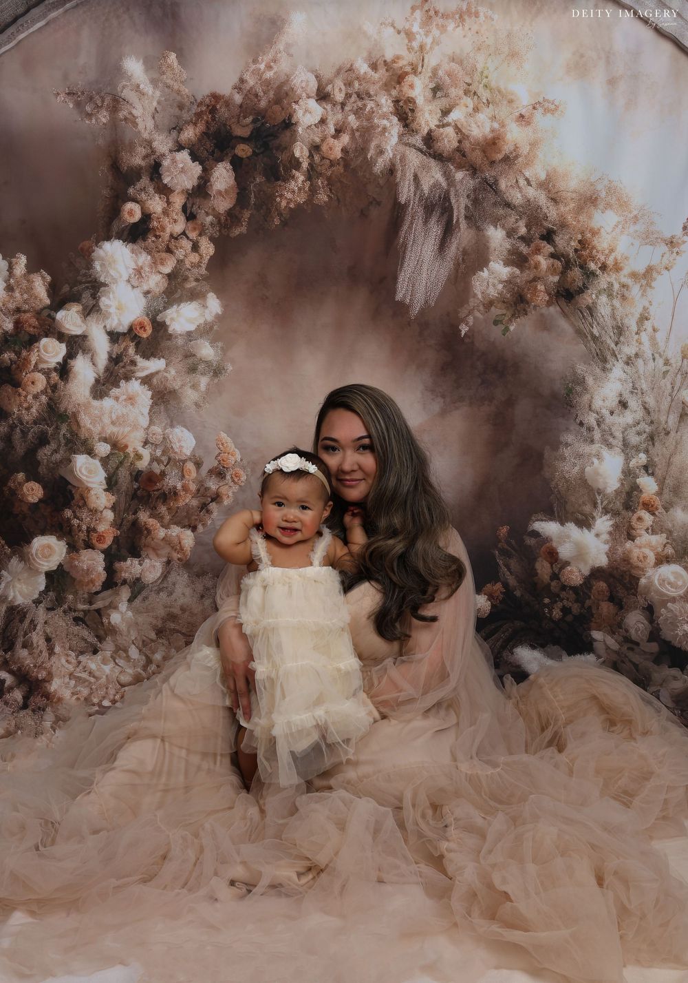 Mother and baby in flowing white dresses pose on floor with romantic floral backdrop in dreamy studio setting.