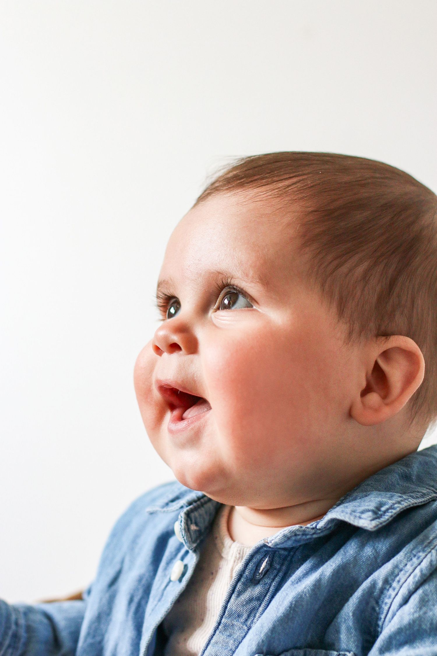 Baby wearing blue denim jacket gazes upward with a joyful expression against white background, during Sitter photoshoot based in Nottingham