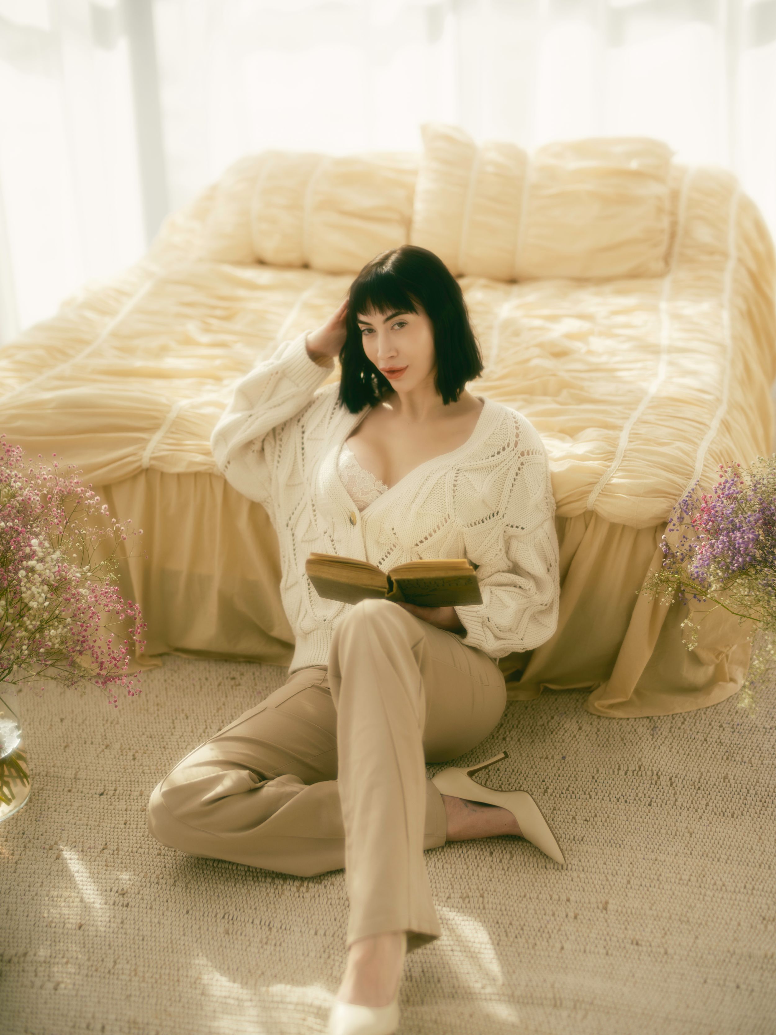 Dreamy vintage-style portrait in soft light shows someone reading a book while sitting on carpet beside a couch with flowers.