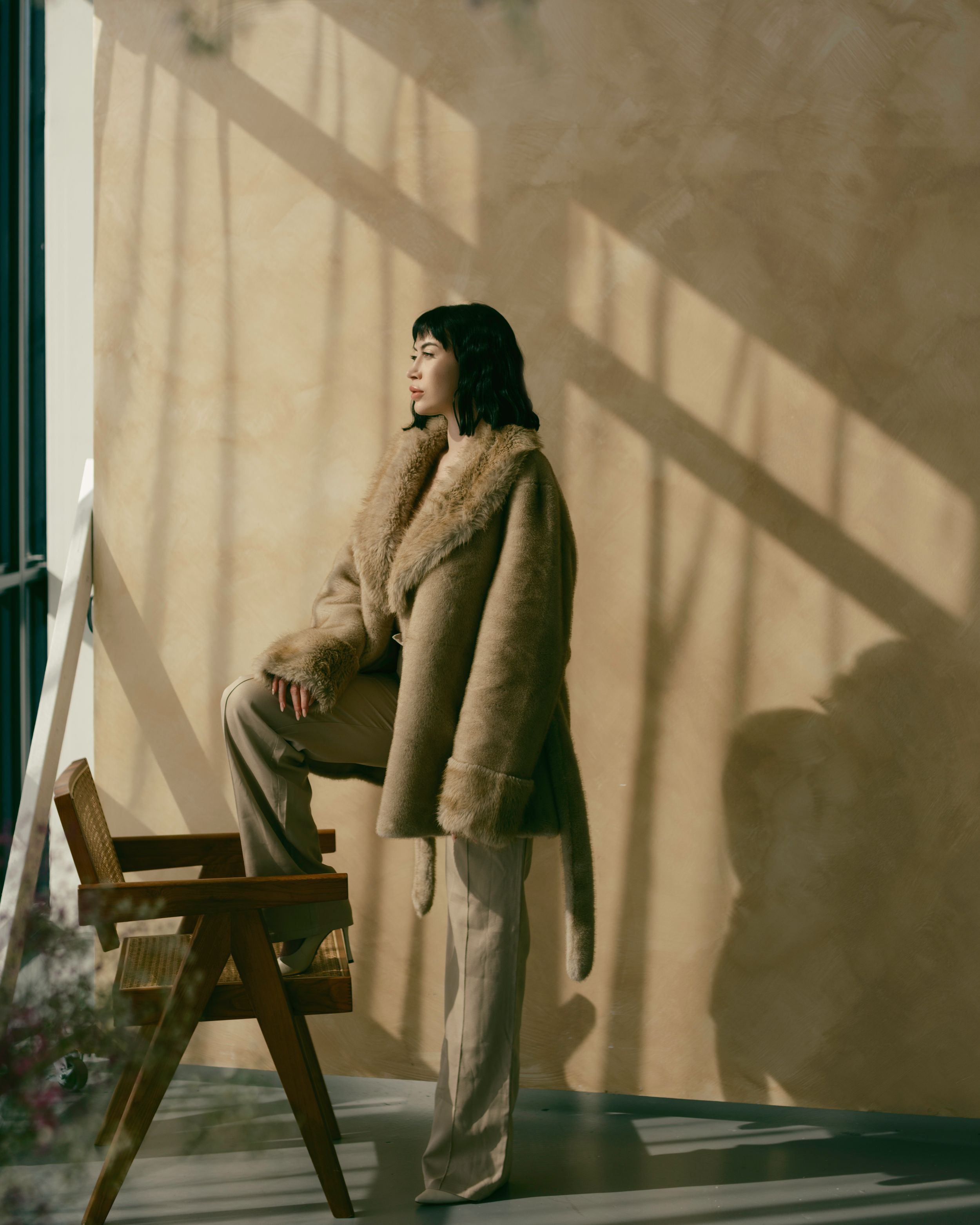 Person in a beige fur coat and wide-leg pants stands near a window, casting dramatic shadows on a bright wall.