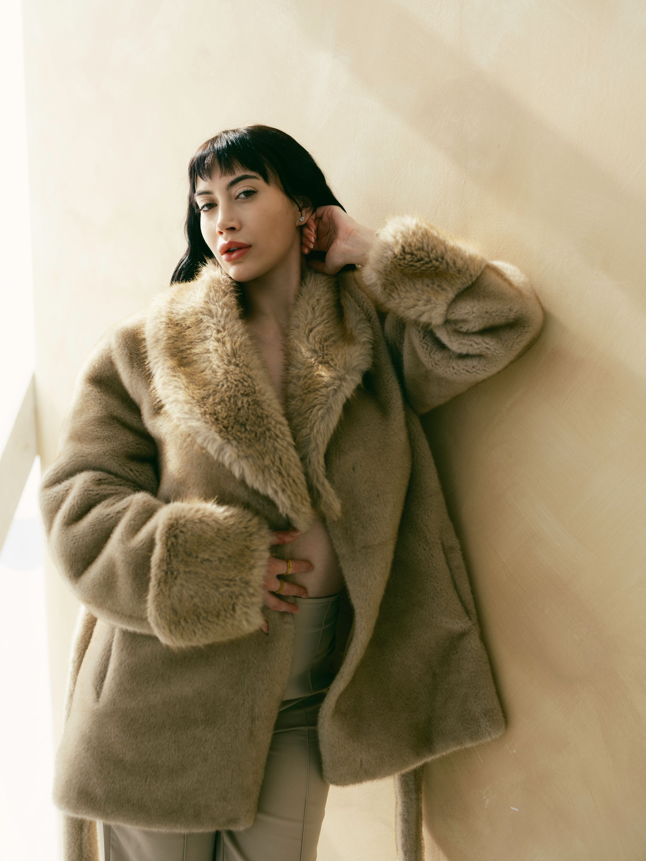 Stylish woman poses in luxurious beige faux fur winter coat against neutral background.