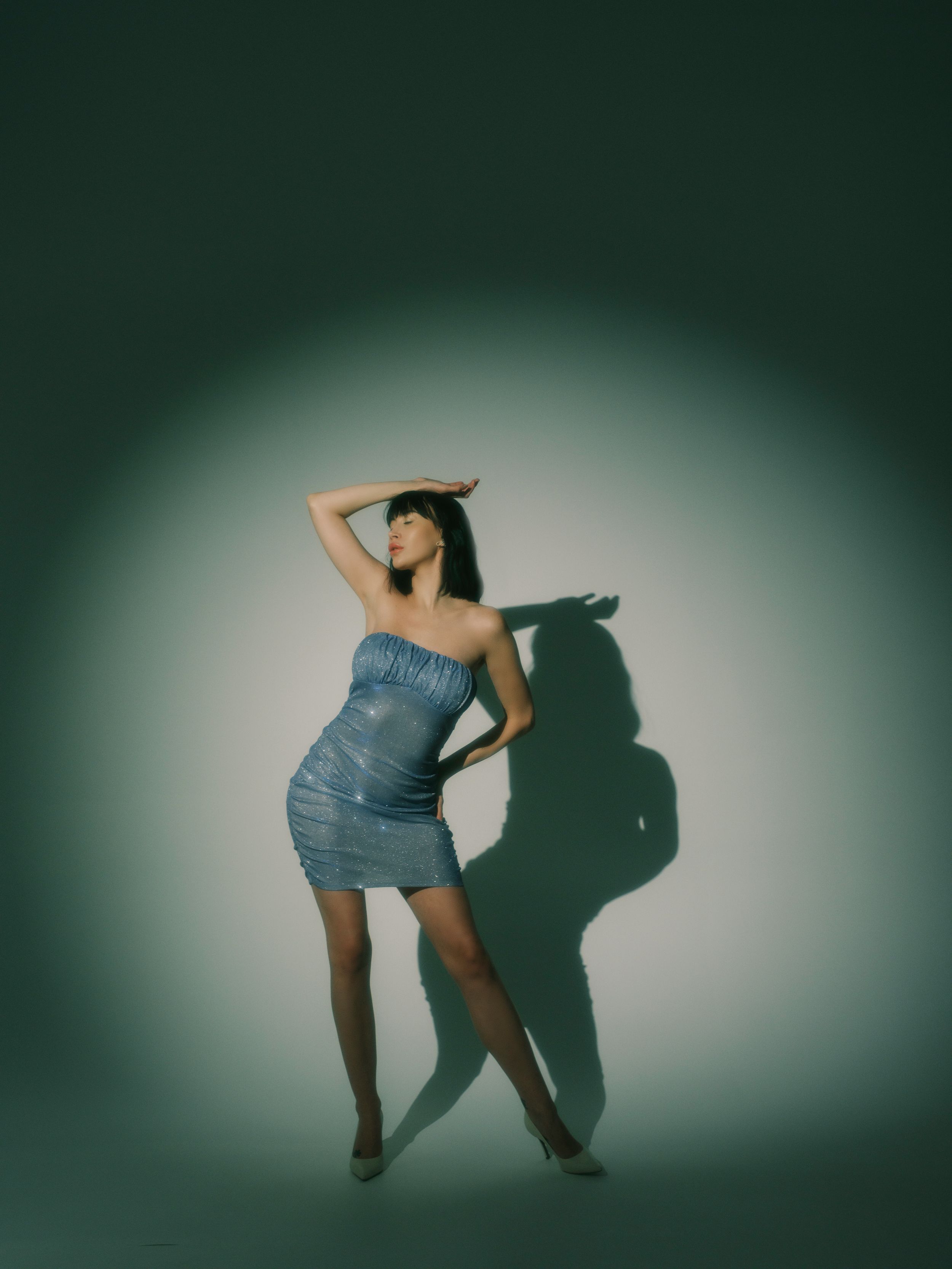 A graceful figure poses in a blue denim strapless dress against a dark green background with dramatic lighting and shadow.