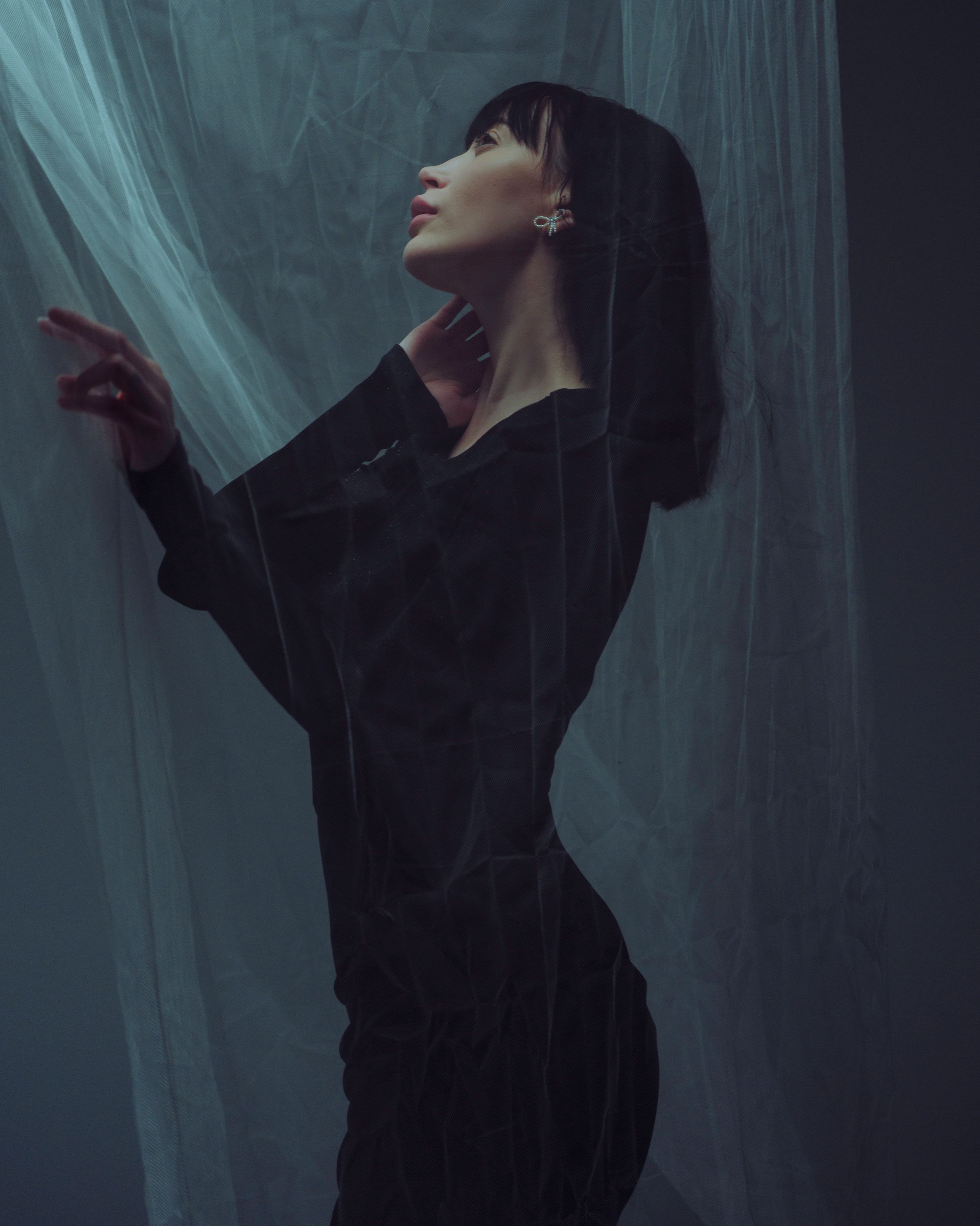 Artistic portrait of a silhouette in black clothing against a teal backdrop with dramatic lighting and flowing fabric.