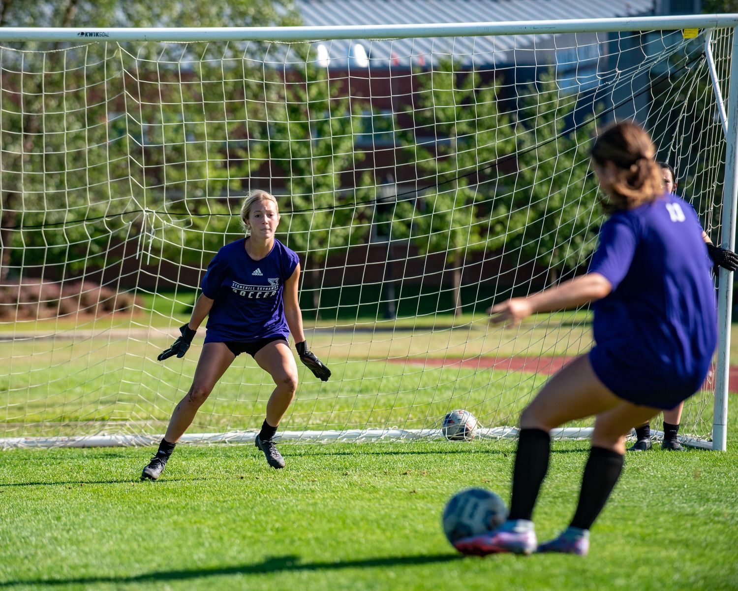 Female soccer goalkeeper in purple jersey preparing to block a kick during practice.