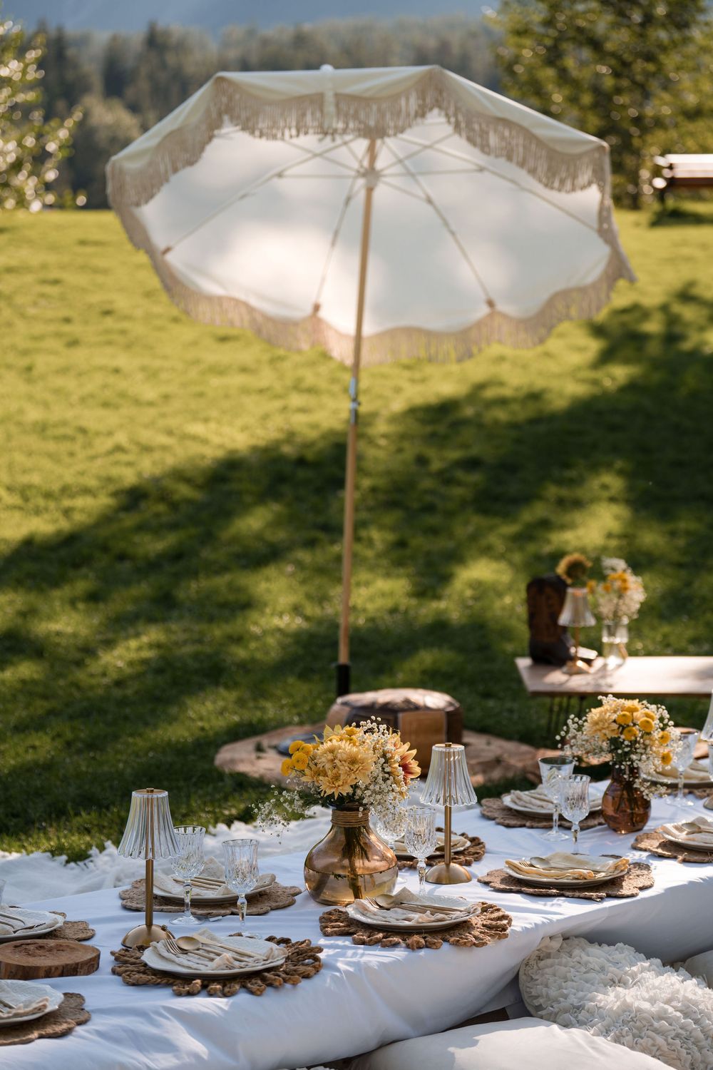 Elegant outdoor picnic table with yellow flowers, gold candles, wicker placemats, and a fringed white umbrella on green lawn.