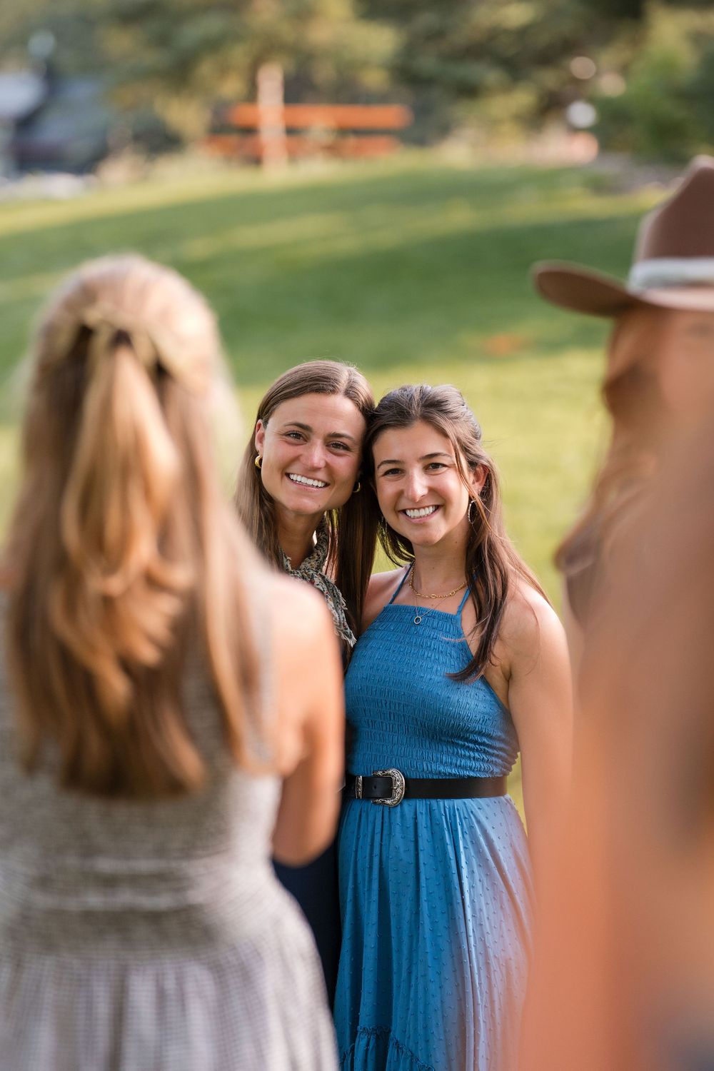 Two smiling young women pose together outdoors at a sunny gathering, wearing stylish summer dresses.