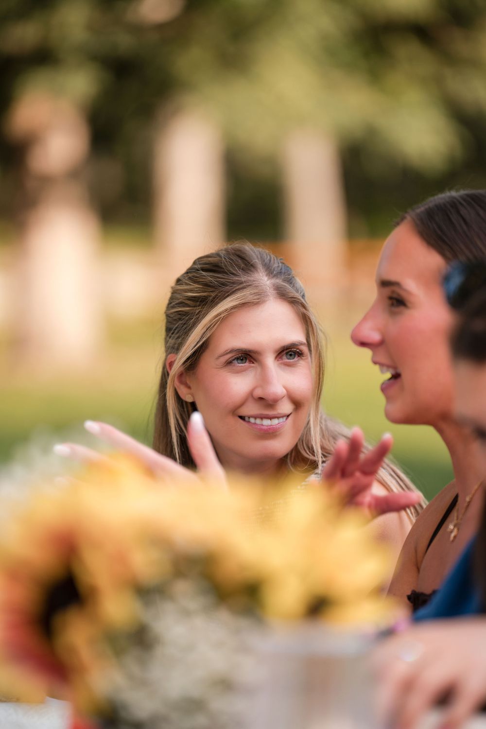 Two women smiling and conversing outdoors, with yellow flowers in the foreground and blurred greenery behind.