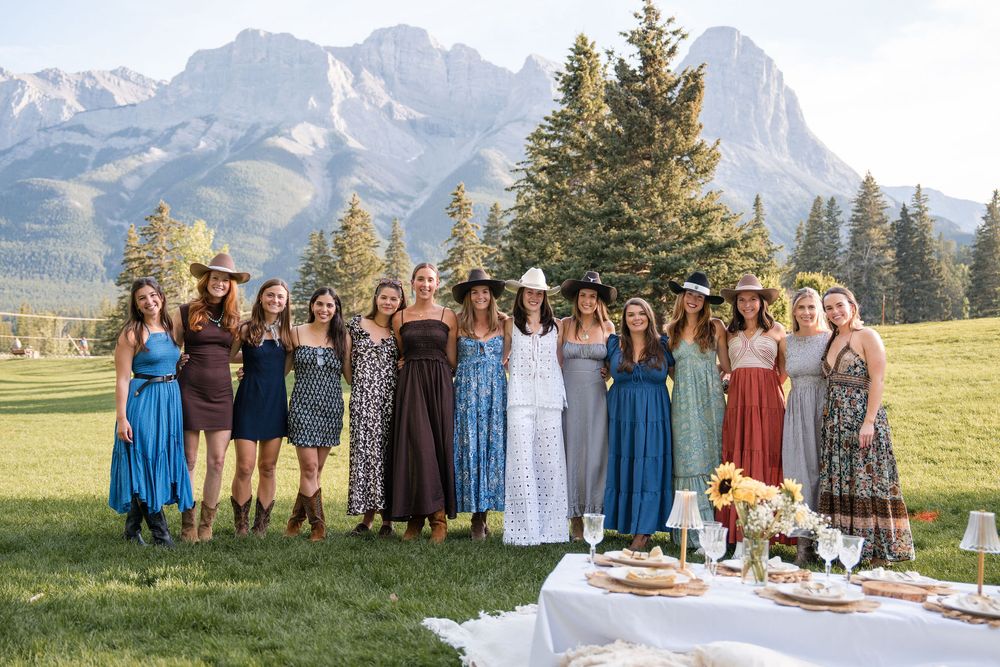 Group of women in boho dresses posing outdoors with mountain and evergreen tree backdrop at a garden party.