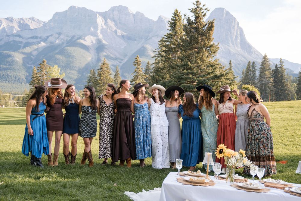 A group of women in colorful summer dresses laugh together on a green lawn with mountain scenery in Canmore, Alberta.