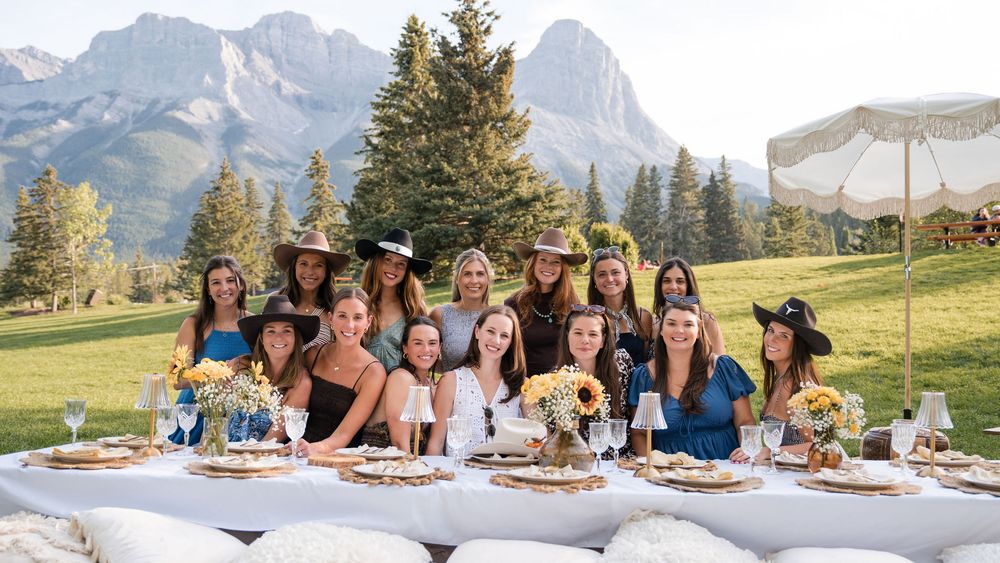 Group of women posing at an outdoor mountain dining table with sunflowers and scenic Rocky Mountain backdrop.