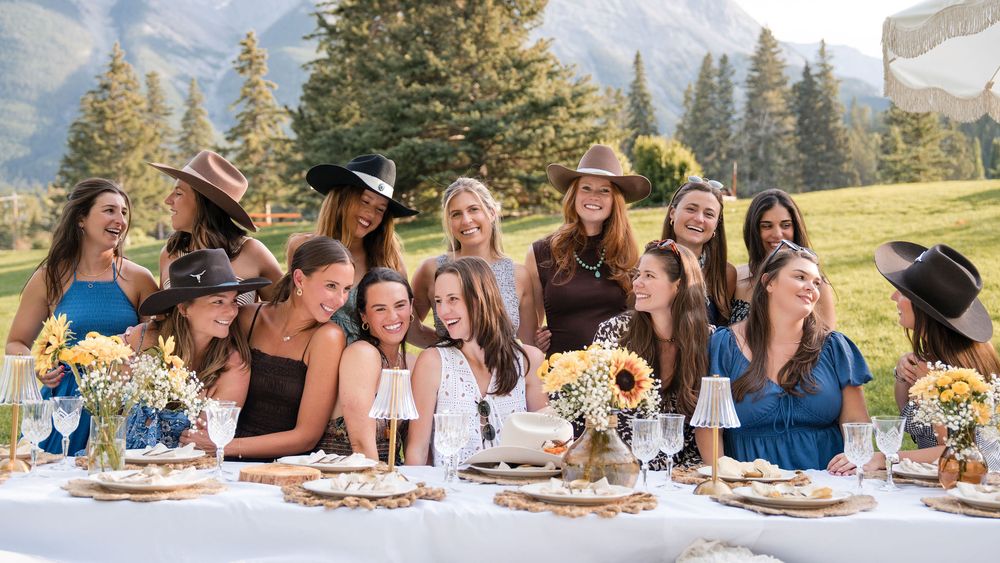 Group of women in cowboy hats celebrating at an outdoor rustic table with sunflowers and mountain views.