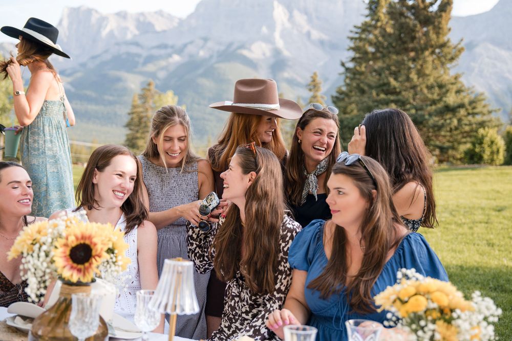 Women laughing and celebrating outdoors at a mountain wedding table with sunflowers and scenic peaks in background.