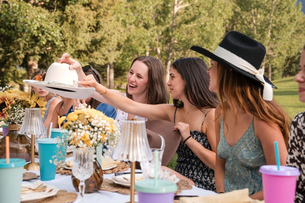 Three women laughing and sharing a white hat at an outdoor garden party table with colorful flowers and drinks.