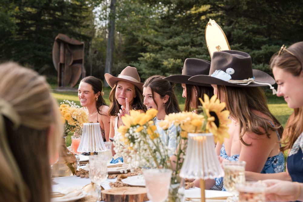 Women in cowboy hats gathered around an outdoor table decorated with sunflowers at a garden party.