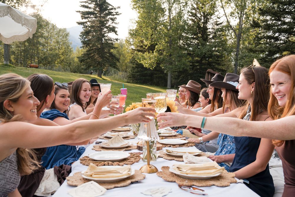 Women toasting drinks at an elegant outdoor dining table surrounded by lush green trees.