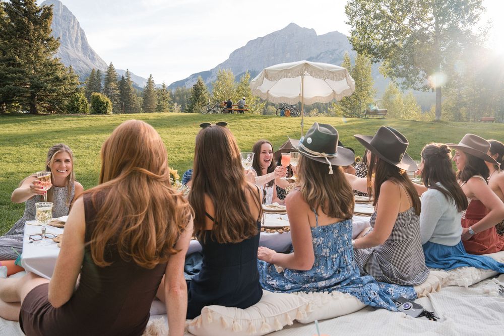 Group of women sitting on grass toasting drinks with mountain views in background.