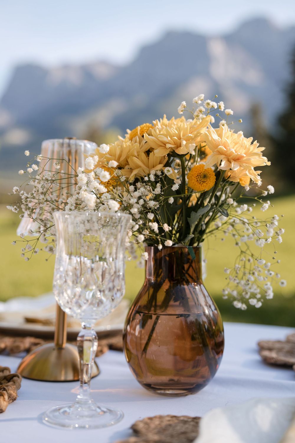 Yellow chrysanthemums and baby's breath in a brown glass vase on an outdoor table with mountain views.