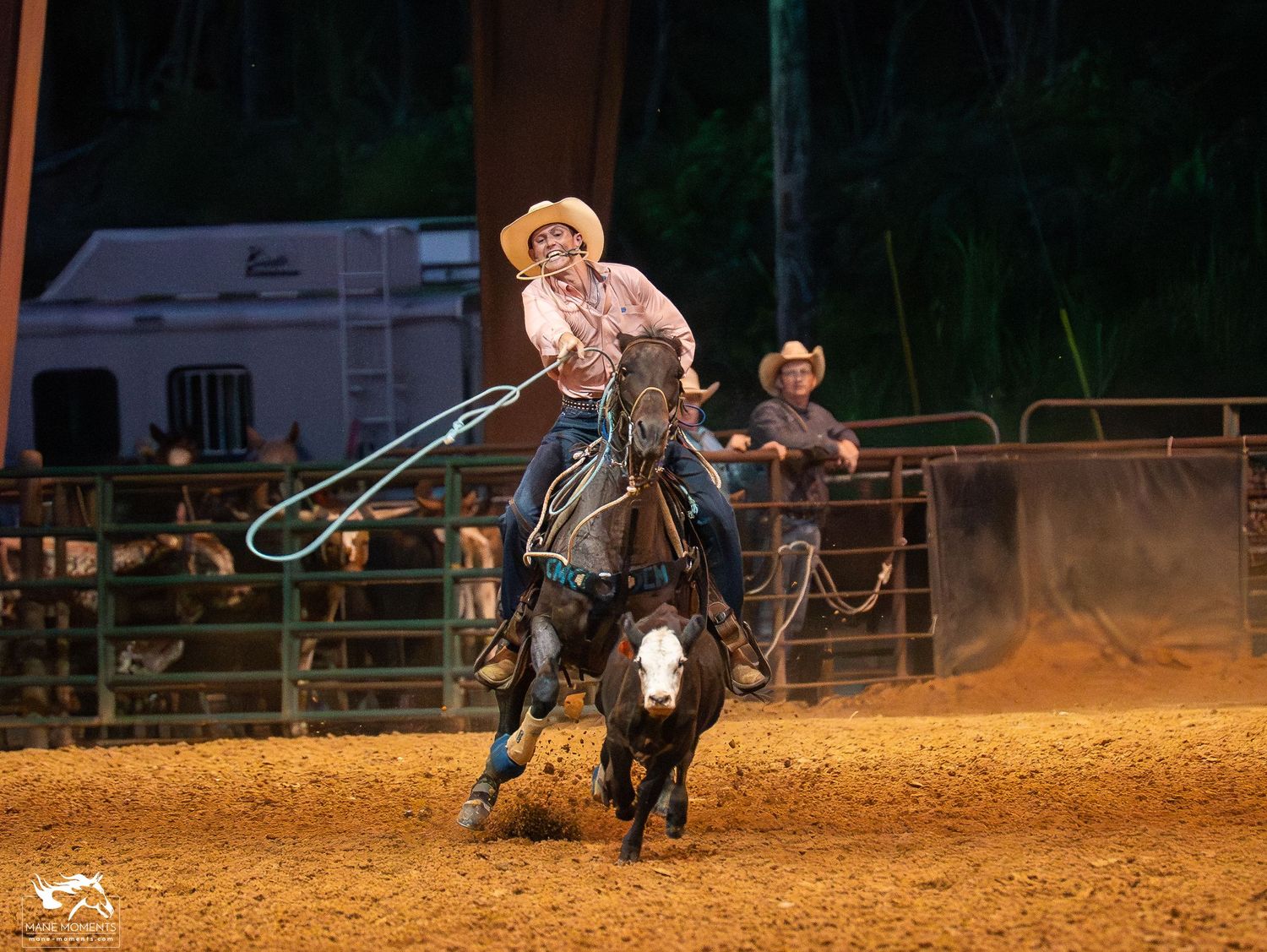 Western rodeo competition featuring roping action in a dusty arena at night with competitors and spectators visible.