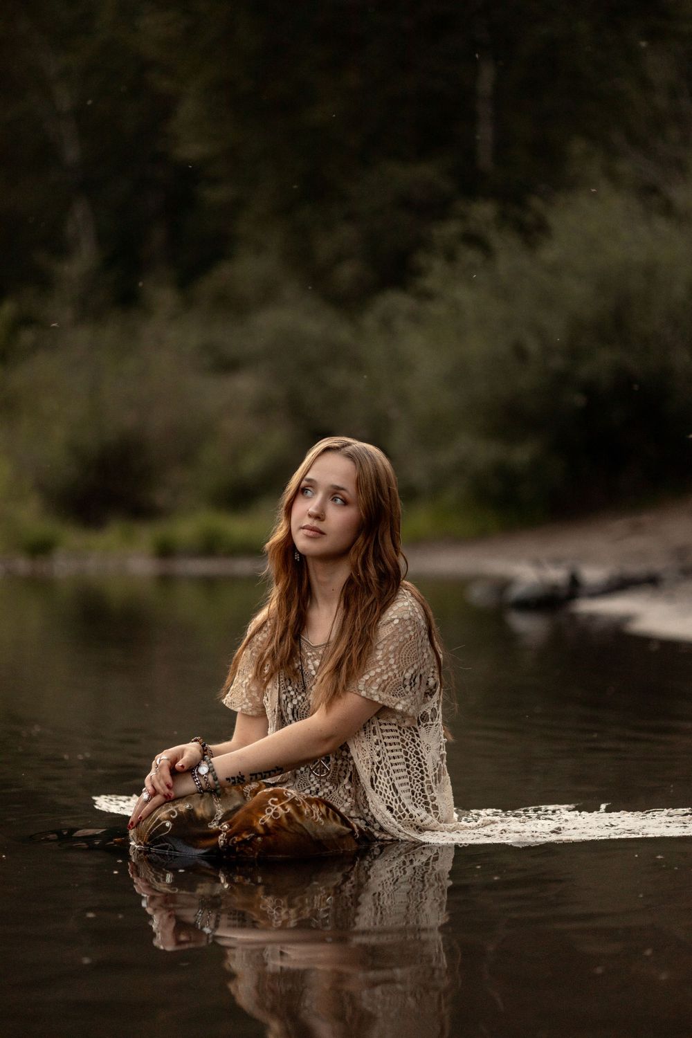 A person in a lace dress sits in shallow river water creating a dreamy ethereal mood during a photography session.