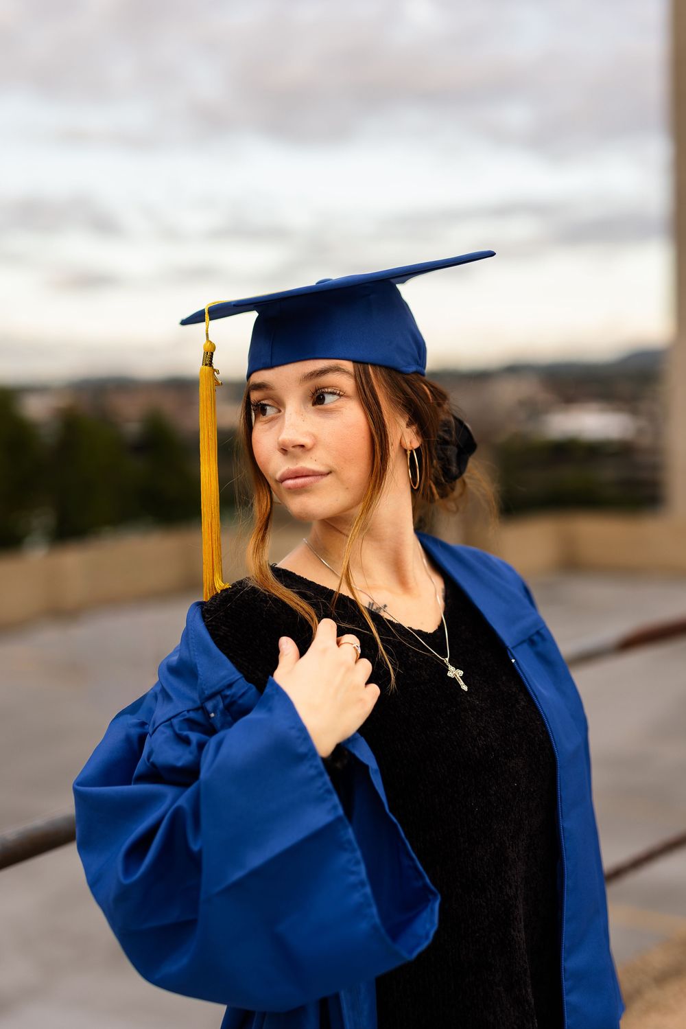 Young woman in blue graduation cap and gown poses outdoors at dusk, looking thoughtfully to the side.