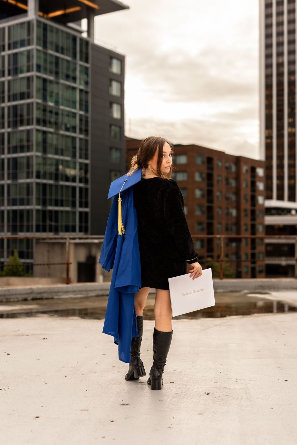 Young woman in blue graduation cap and gown holding diploma, walking outdoors with city buildings in background.