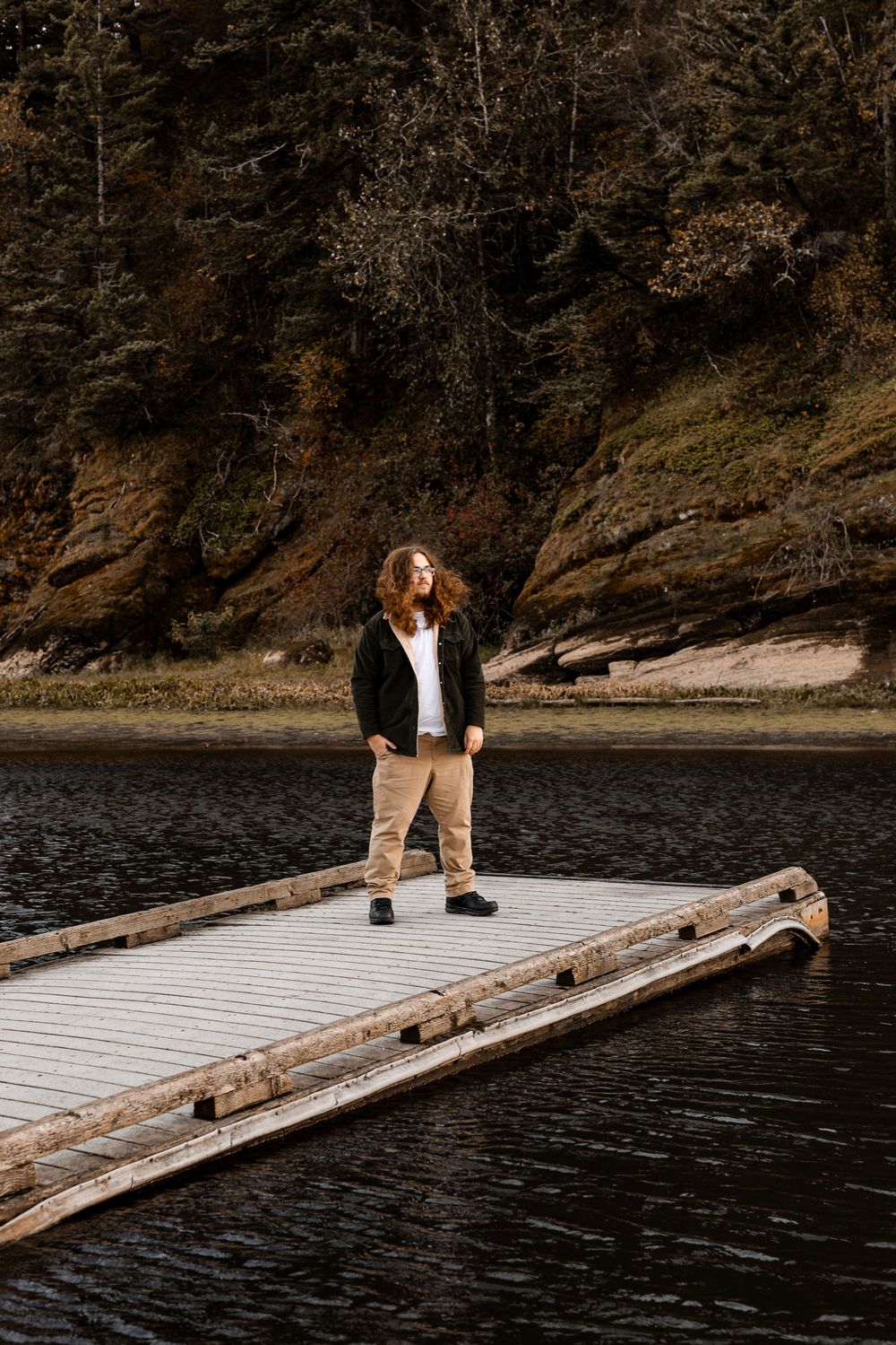 Person with long hair standing on a floating dock surrounded by dark water with rocky cliffs and trees in background.