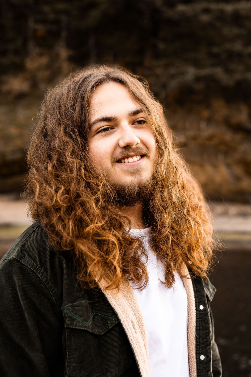Young man with long curly auburn hair and beard smiling outdoors, wearing a dark jacket over a white shirt.