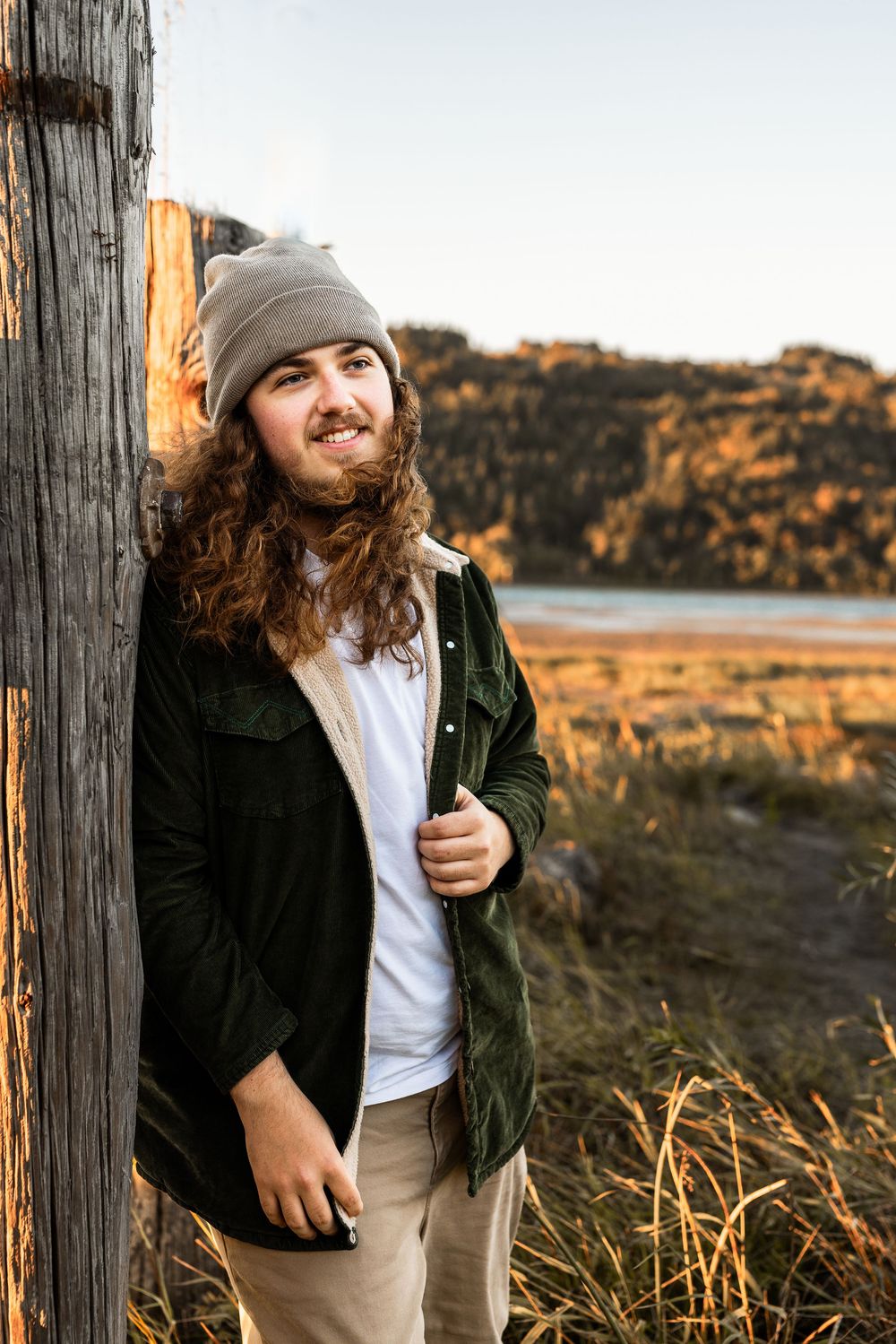Young man with long curly hair and gray beanie leans against wooden post in golden outdoor landscape.