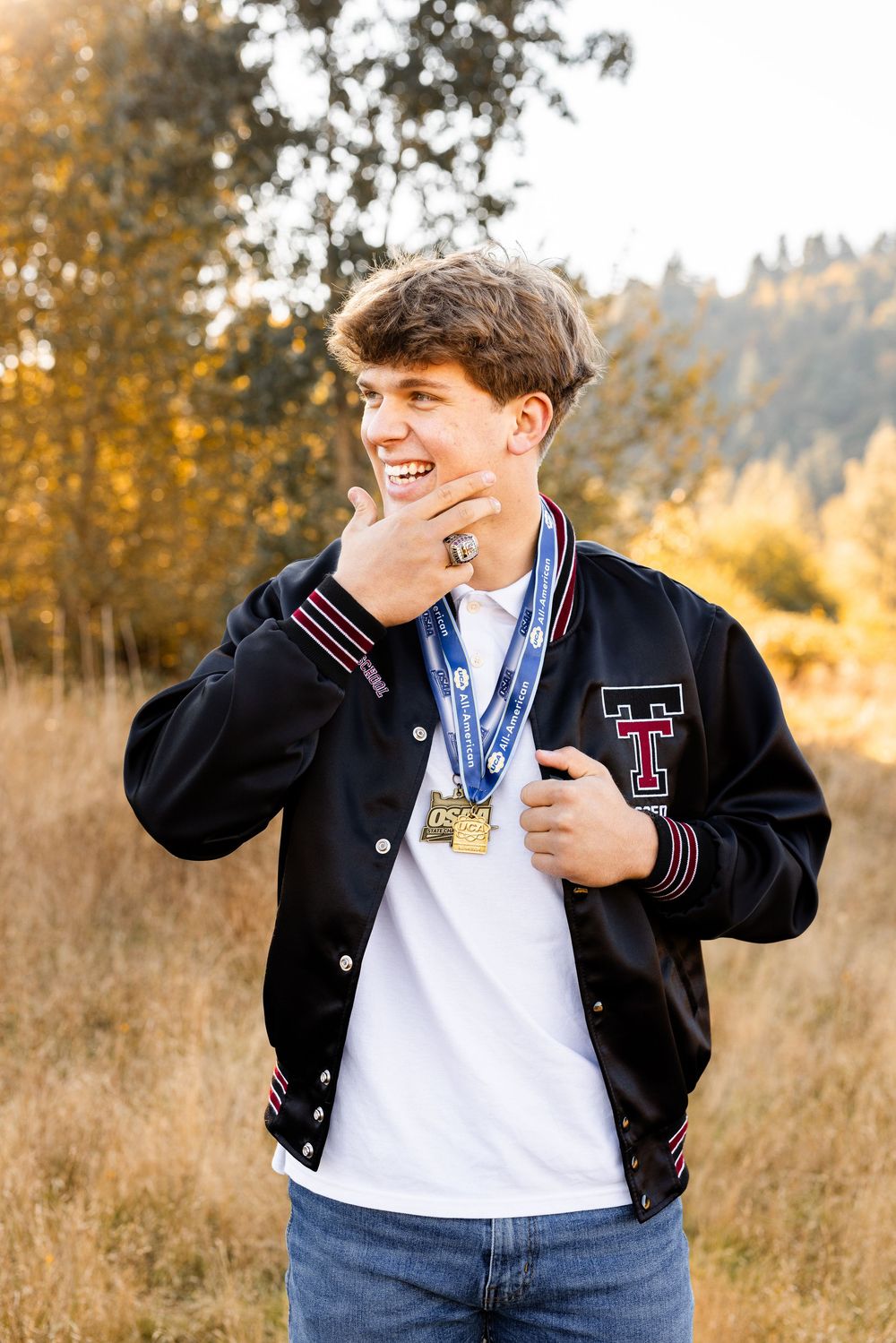 Smiling teenage boy wearing a black varsity jacket with medals outdoors in a golden autumn field.