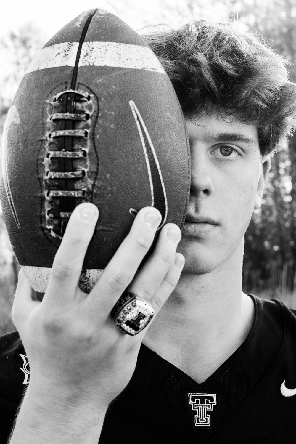 Black and white photo of a young male football player holding a football over half his face, wearing a ring and Nike jersey.