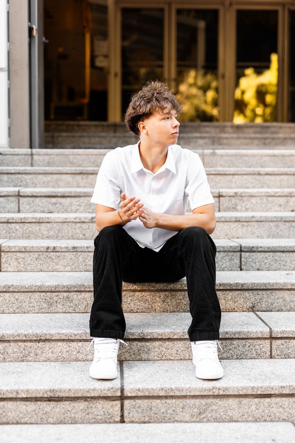 Young man in white shirt and black pants sitting on outdoor stone steps, looking away thoughtfully.