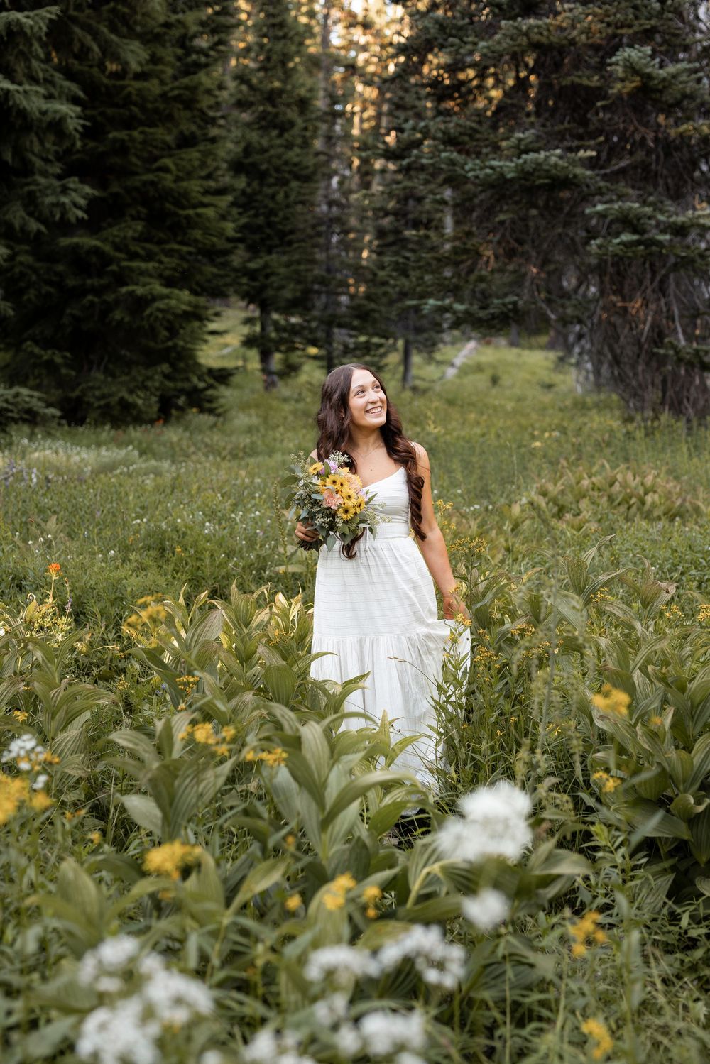 Smiling bride in white dress holding yellow wildflowers in a lush mountain meadow surrounded by evergreen trees.