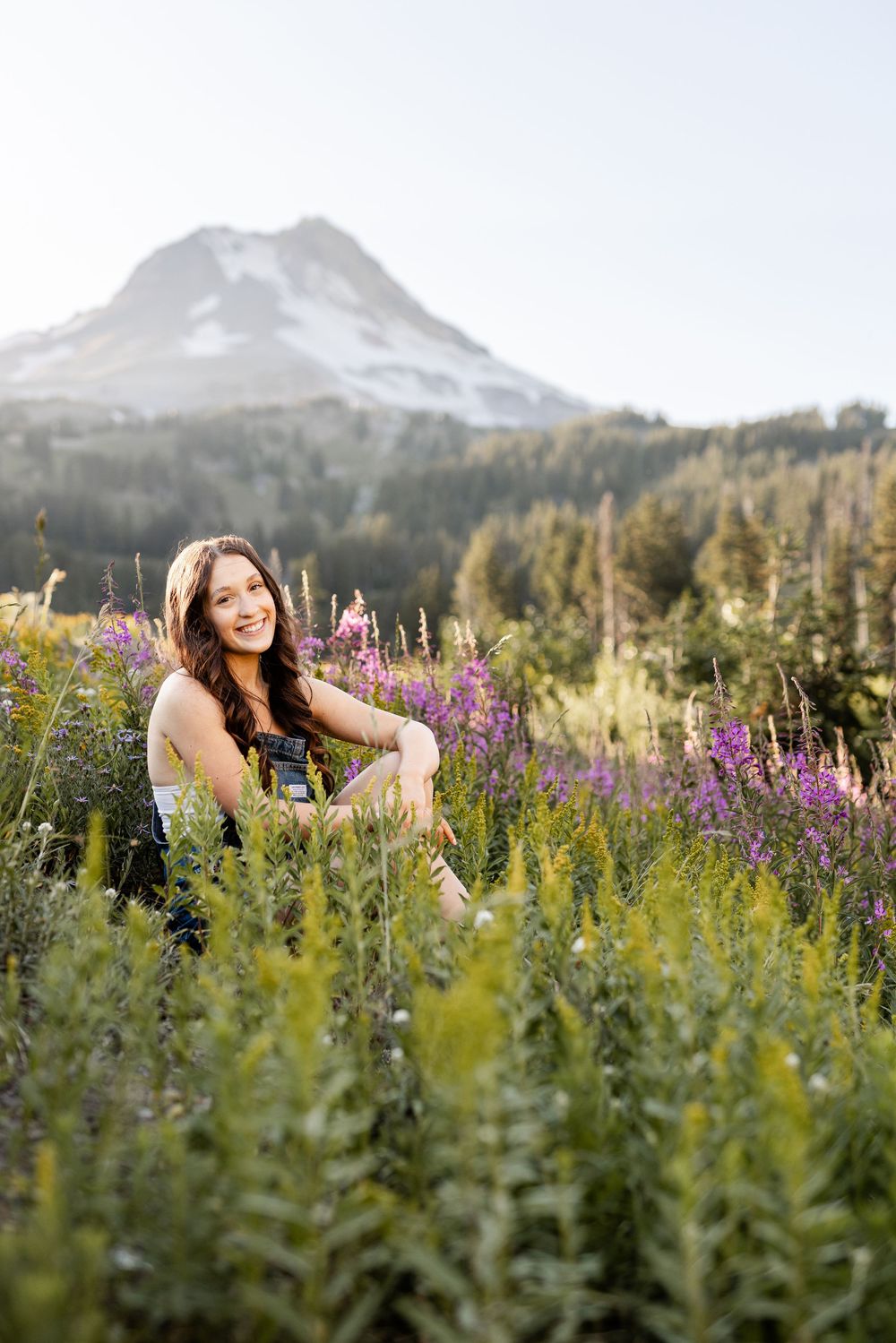 Young woman sitting in a wildflower meadow with purple blooms, smiling, with a snow-capped mountain in the background.