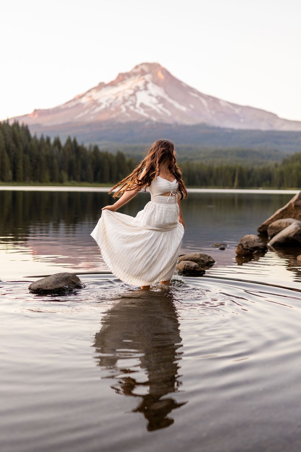 A scenic mountain lake view with a person in a flowing white dress standing on rocks at Trillium Lake near Mount Hood.