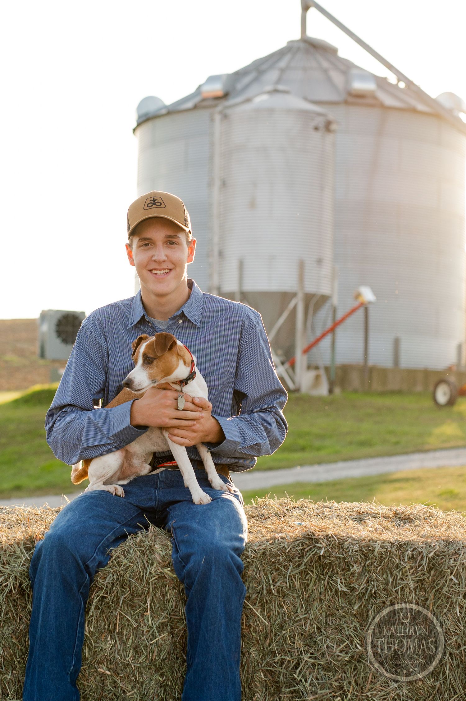 Person sits on hay bale holding a small dog with a grain silo in the background during golden hour.