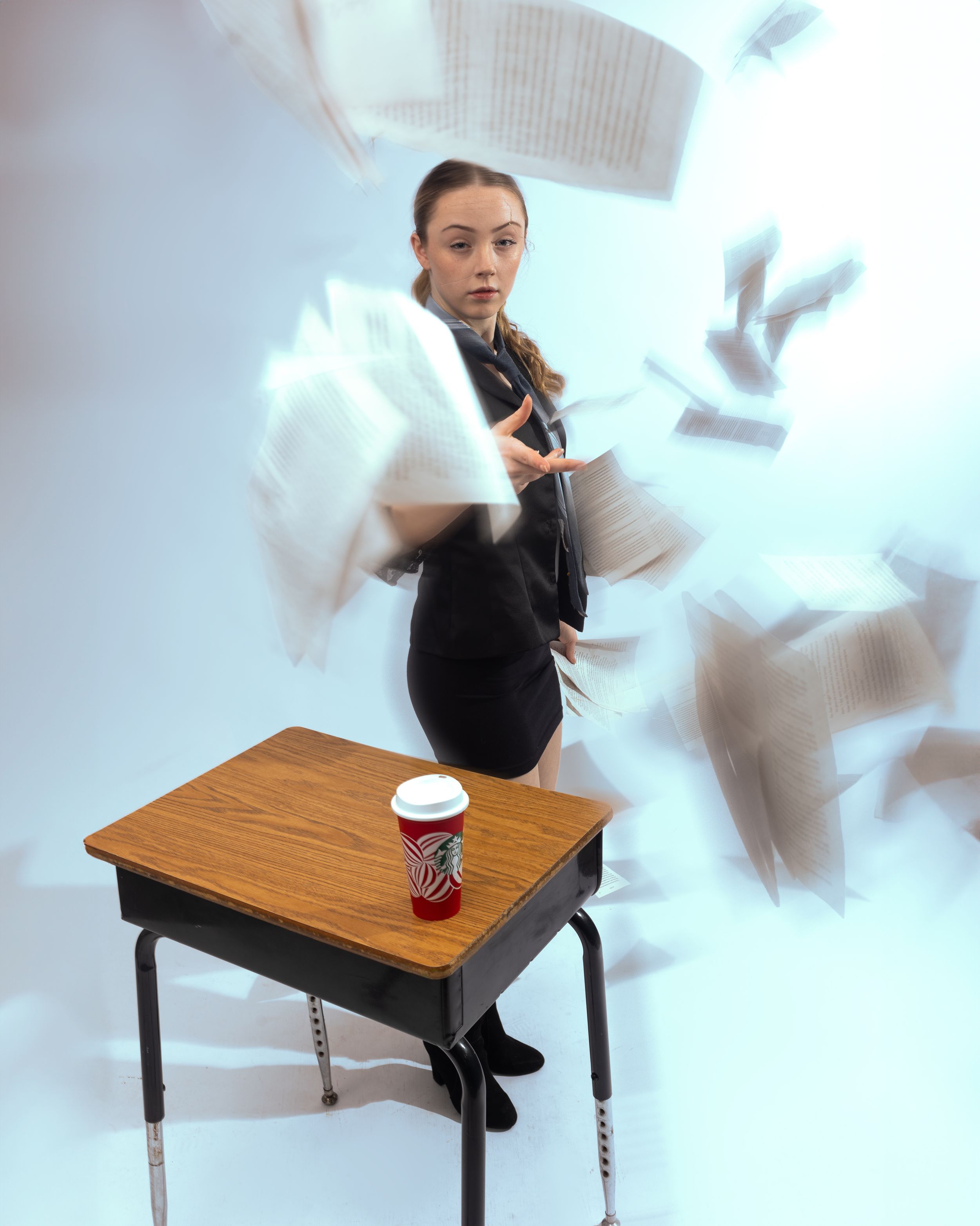 Someone in a black outfit stands on a desk with papers flying through the air and a coffee cup on the surface.